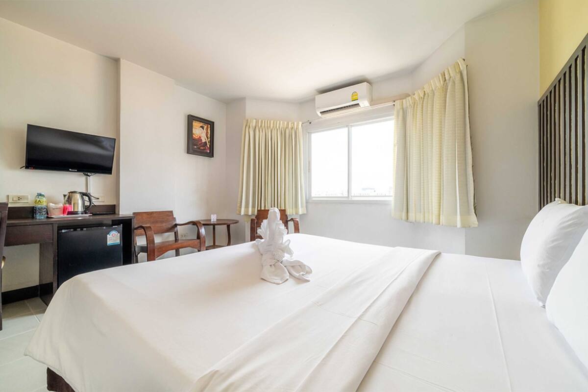 A hotel room with a bed, a desk with a television, a chair, and an air conditioning unit.