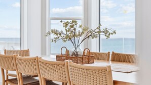 A dining area with wicker chairs and a table, a wicker basket with a vase, and a view of the sea through large windows.