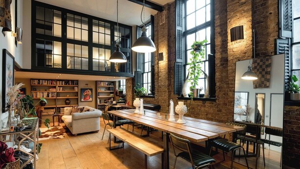 Een ruime loft met onbehandelde bakstenen muren, grote ramen, een houten eettafel en een gezellige zithoek.