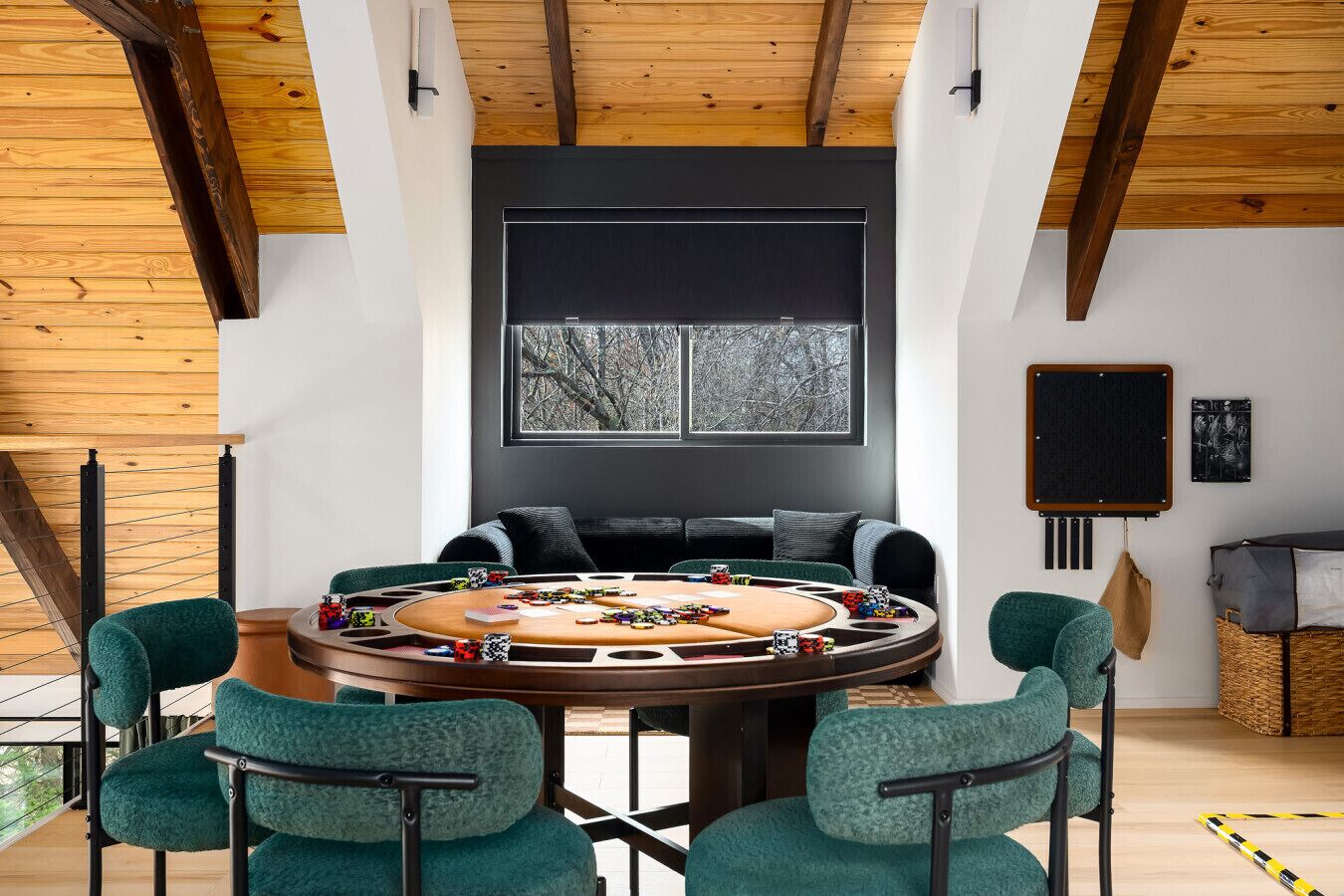 Une pièce moderne avec un plafond en bois, une table ronde sur laquelle sont disposés des jetons de poker, des chaises vertes, un canapé noir et une grande fenêtre donnant sur des arbres.