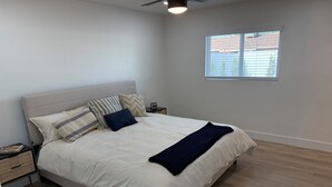 Una cama bien tendida con un cabecero blanco, varios almohadones y un plaid oscuro. Hay una mesita de noche con una lámpara y un libro. El cuarto tiene piso de madera clara y una ventana con persianas.