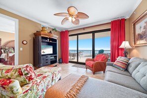 Sala de estar con sofá, sillón y televisor. Balcón con cortinas rojas y vistas al exterior.