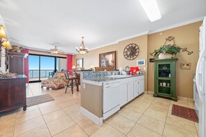 A spacious kitchen with a large island, a dining area with a chair and table, and a view through the glass door. - Beachfront Condo w/ Huge Balcony, Pools, Sauna & Gulf Views | BEACHBALL PROPERTIES (Orange Beach)