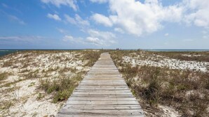 Una pasarela de madera que lleva a la playa, rodeada de dunas de arena y vegetación escasa.