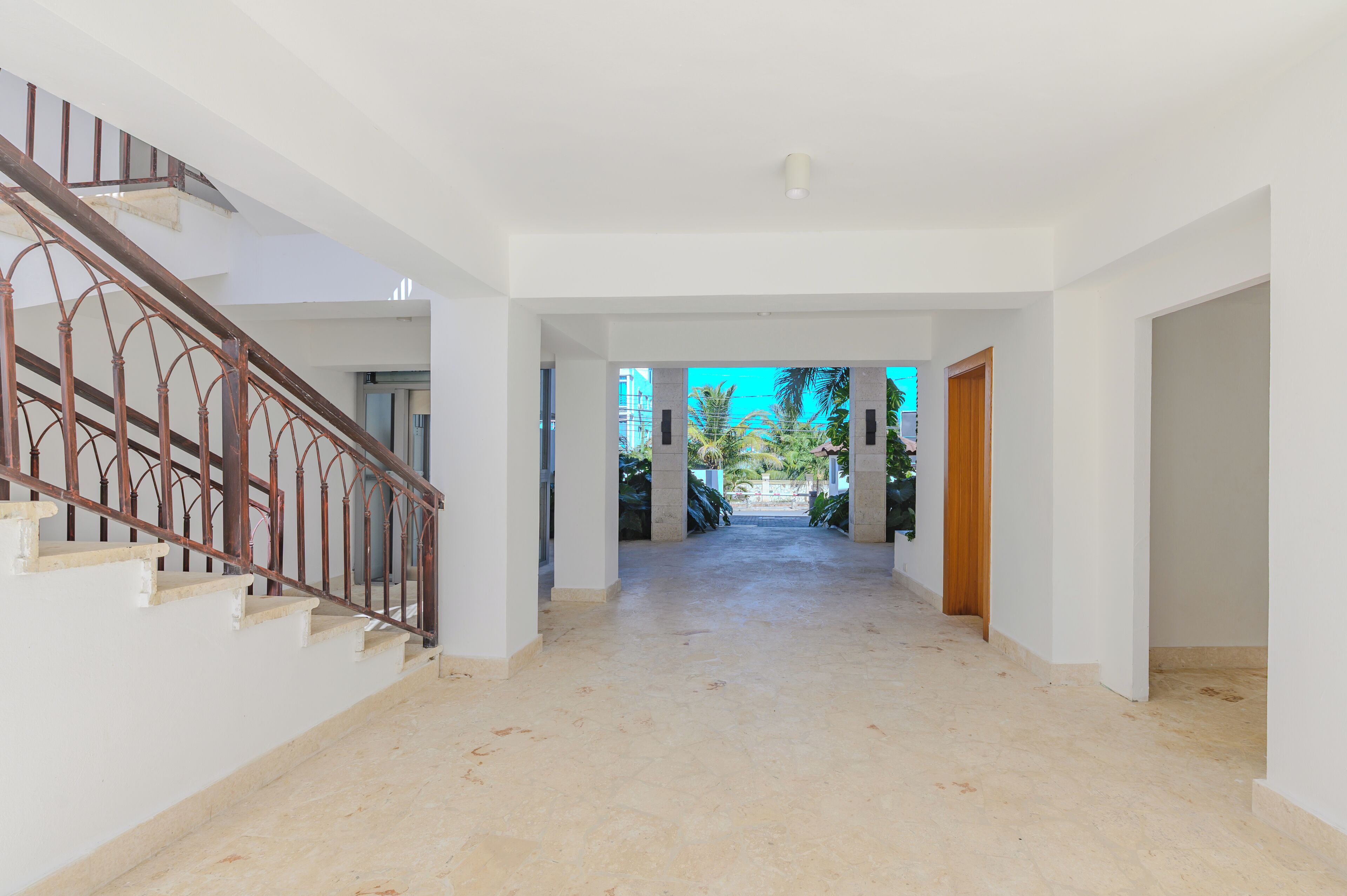 A spacious interior with a staircase leading to upper floors, a hallway with a view of greenery outside, and a door on the right.