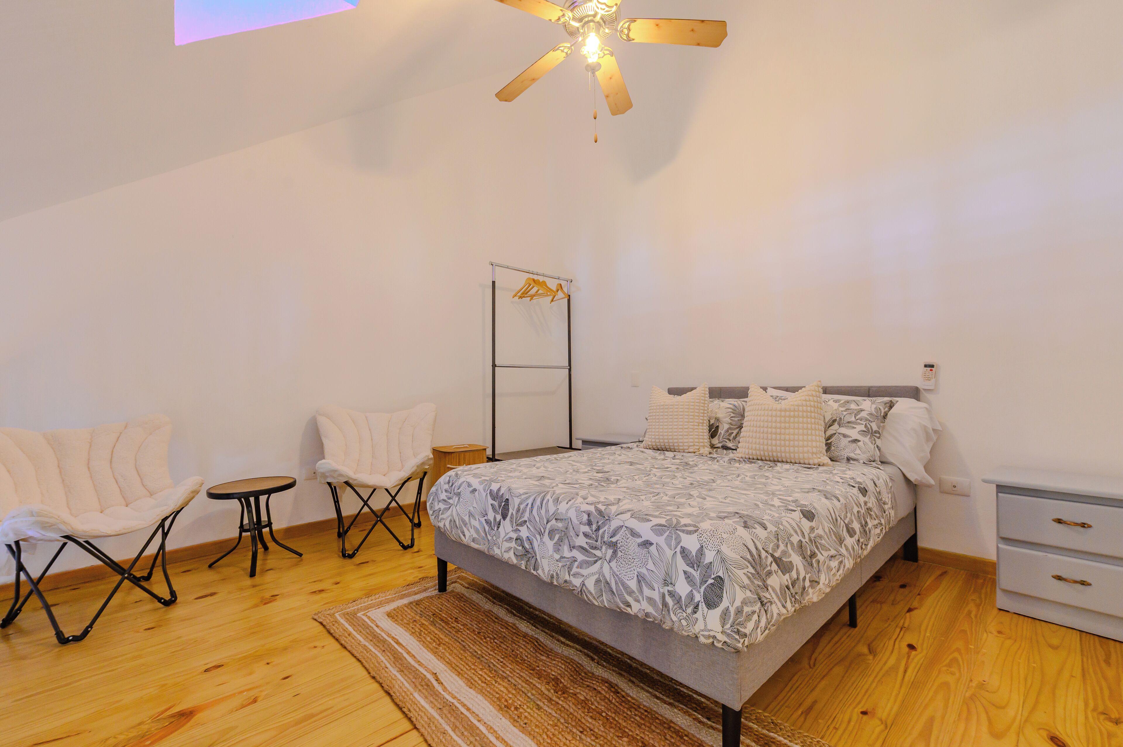 A bedroom with a bed, two chairs, a small table, a ceiling fan, and a nightstand.
