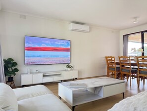A modern living room with a flat-screen TV, white furniture, a dining area with wooden chairs, and a large window with curtains. - 8 BR Retreat - 2 Full Houses on Property (Frankston)