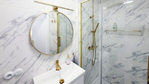A bathroom with a marble wall, a round mirror with a gold frame, a gold faucet, and a glass-enclosed shower with gold fixtures. - Cozy 1-bedroom studio in amazing Katowice, 24h check-in (Katowice)