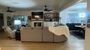 A spacious living room with a large sectional sofa, a dining area with a table and chairs, and a kitchen area with a TV and shelves. - Spacious 4-bedroom house in beautiful Bartlesville with AC (Bartlesville)