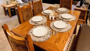 Une salle à manger avec une table en bois dressée pour six personnes, entourée de chaises dotées de dossiers finement ouvragés. On aperçoit une cheminée au fond de la pièce, et un téléviseur est fixé au mur.