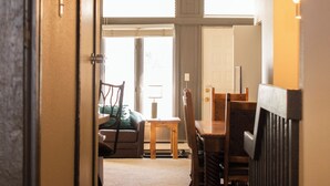 Un couloir mène à une salle à manger équipée d’une table et de chaises, d’un canapé et d’une fenêtre avec des stores.