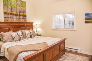 A bedroom with a wooden bed, a nightstand with a lamp, a window with blinds, and a painting on the wall.