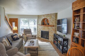 A living room with a stone fireplace, a flat-screen TV, a bookshelf, and a staircase.