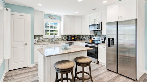 A modern kitchen with white cabinets, a stainless steel refrigerator, and a central island with stools. - Unwind and Relax at Tybee Mist | Heated Pool Access (Tybee Island)