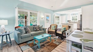 A living room with a sofa, coffee table, and chairs, a dining table with placemats, and a kitchen area in the background. - Unwind and Relax at Tybee Mist | Heated Pool Access (Tybee Island)