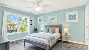 A bedroom with a bed, a ceiling fan, a window with a view of greenery, a bedside table with a lamp, and a rug. - Unwind and Relax at Tybee Mist | Heated Pool Access (Tybee Island)