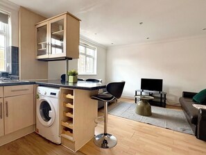 Ein kompakter Wohnbereich mit Küche, Waschmaschine, Barhocker, Fernseher und Sofa.