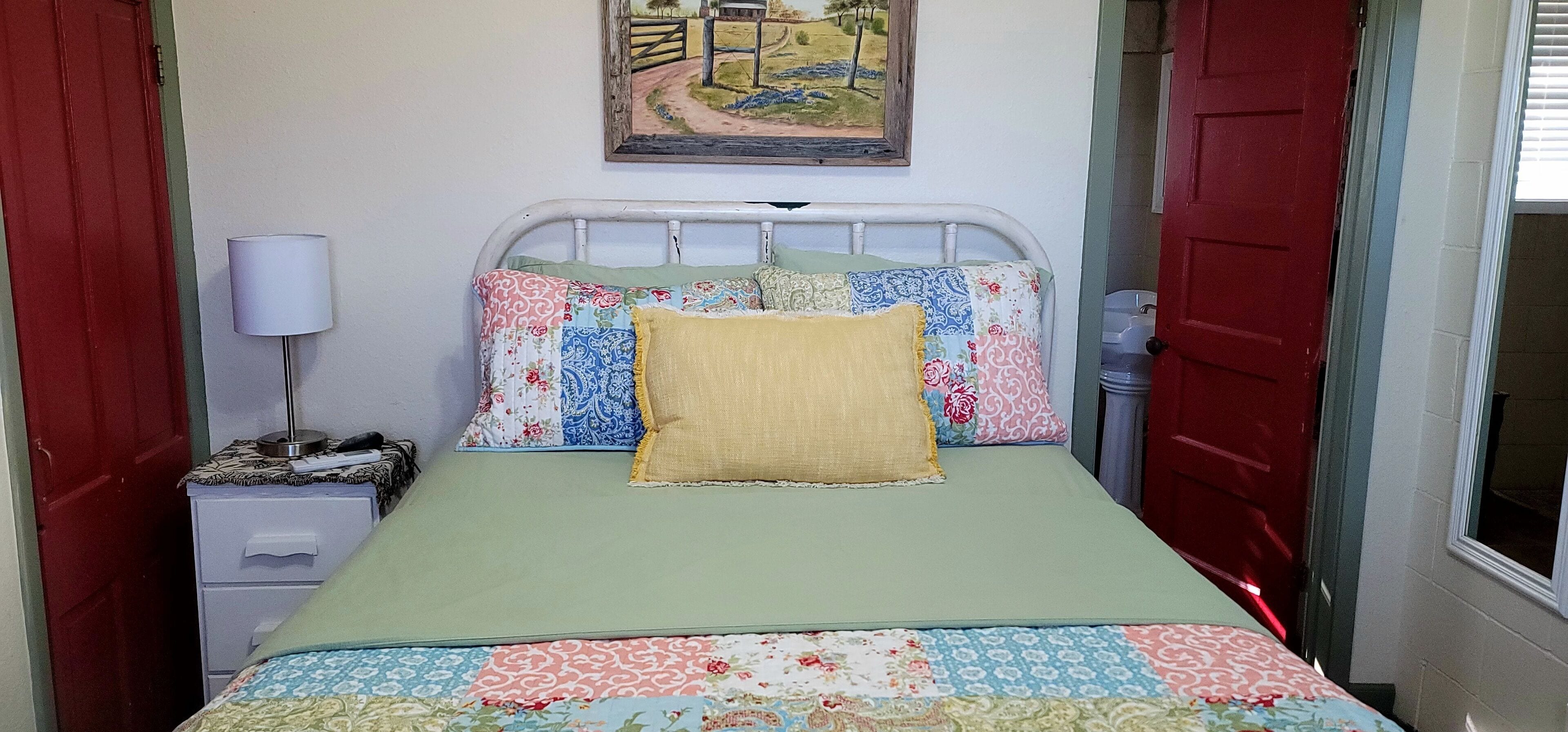 A small, neatly arranged bedroom with a single bed, a bedside table with a lamp, and a painting on the wall.