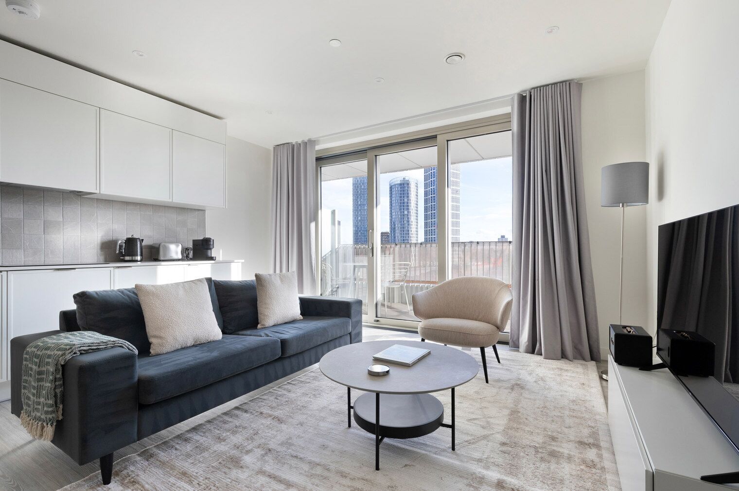 Un salon moderne avec un canapé bleu foncé, une table basse ronde et un fauteuil beige. On y trouve une grande fenêtre avec des rideaux, un téléviseur à écran plat et un coin cuisine équipé de placards et d’appareils électroménagers.