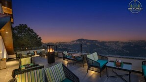 Terraza en la azotea con mobiliario exterior y una chimenea, con vistas a un paisaje urbano al anochecer.