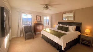 A hotel room with a bed, a chair, a dresser, a nightstand, a ceiling fan, and a large window with a view. - The Susie Pearl Bed & Breakfast (Kelowna)