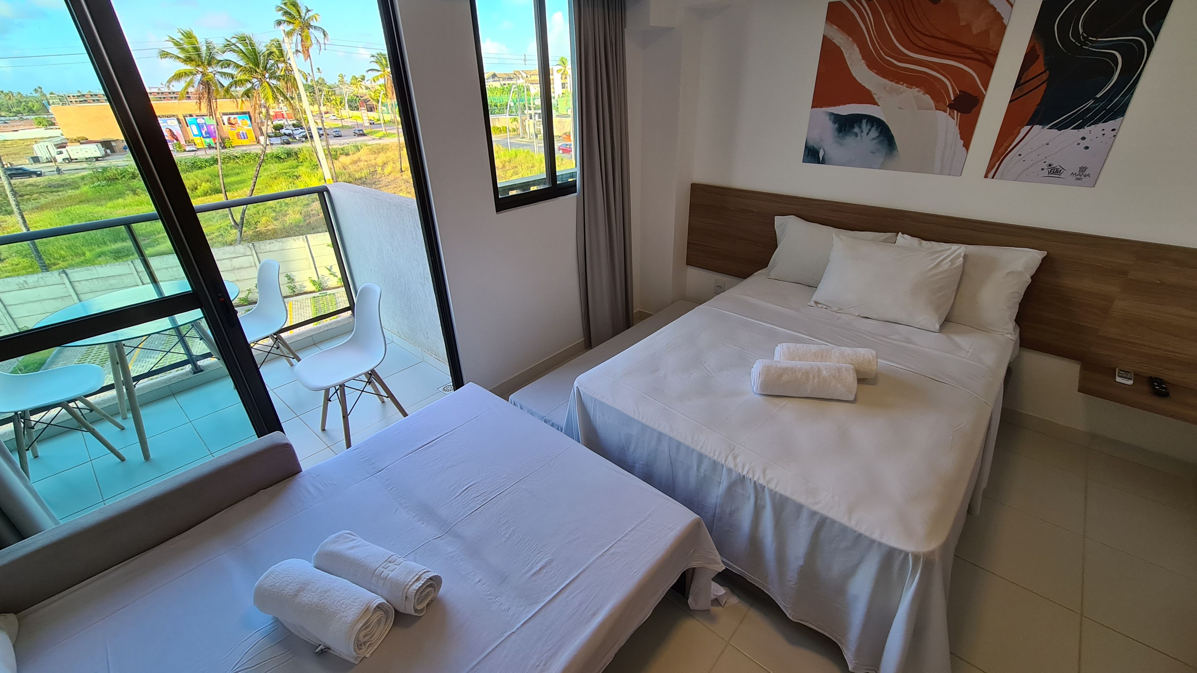 A hotel room with two beds, a balcony with a view of palm trees and a road, and a painting on the wall.