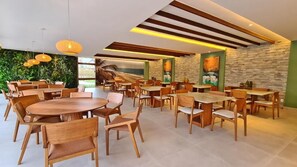 A spacious dining area with multiple wooden tables and chairs, a green wall with a beach view, and a stone wall with artwork. - Mana Beach Experience By Aft (Ipojuca)