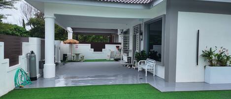 A covered outdoor area with artificial grass, a white bench, a potted plant, and a thatched umbrella.