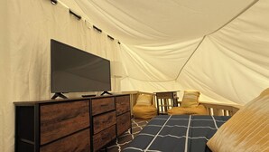Une pièce aménagée comme une tente, avec un téléviseur posé sur une commode en bois, un lit avec une literie à motifs et une petite table entourée de chaises.