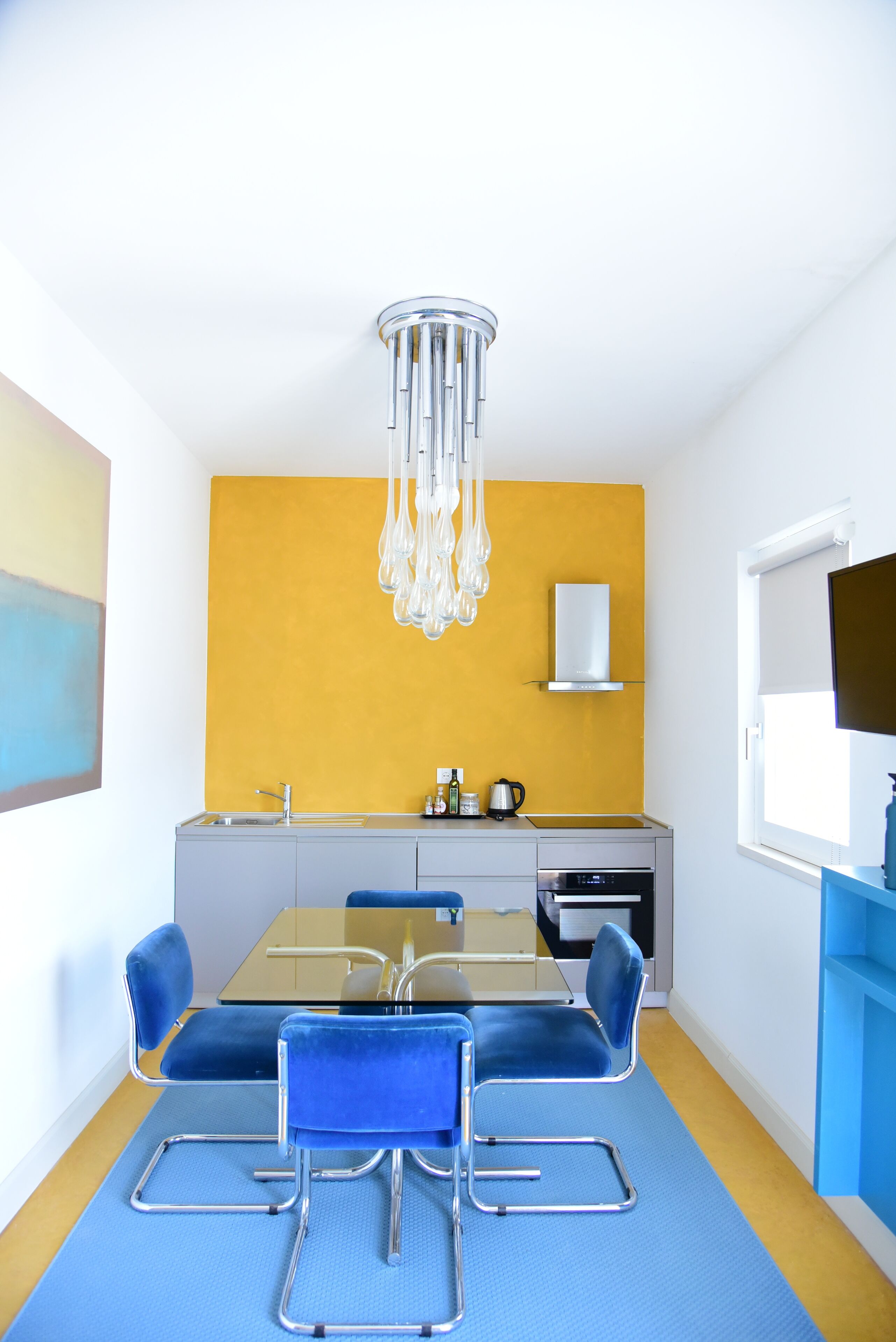 A modern kitchen with a yellow accent wall, a dining table with blue chairs, and a built-in oven.