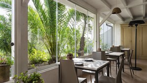 A room with a large window overlooking lush greenery, a dining table with chairs, and a floor lamp.