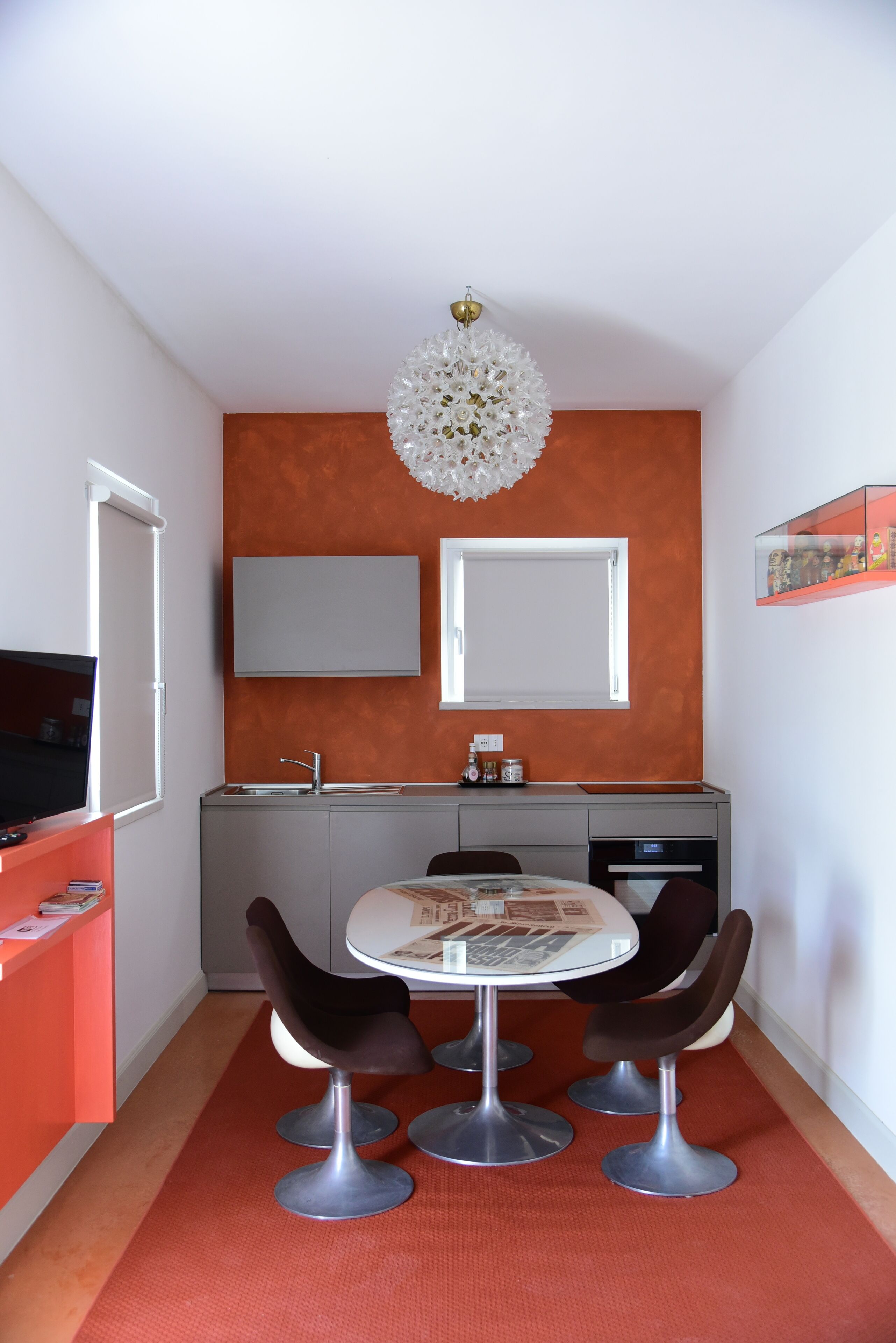 A compact kitchen with a modern design, featuring a white ceiling, an orange accent wall, and a chandelier.
