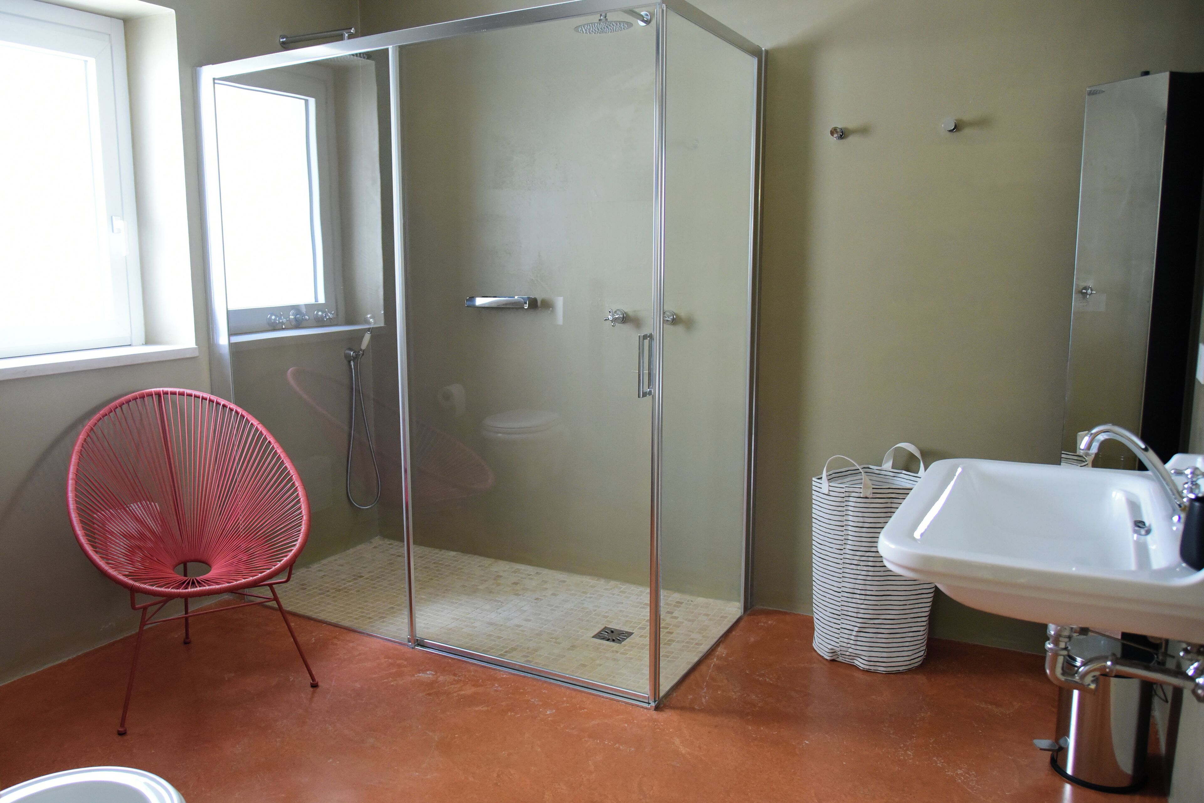 A bathroom with a glass shower enclosure, a red chair, a sink, and a trash bin.