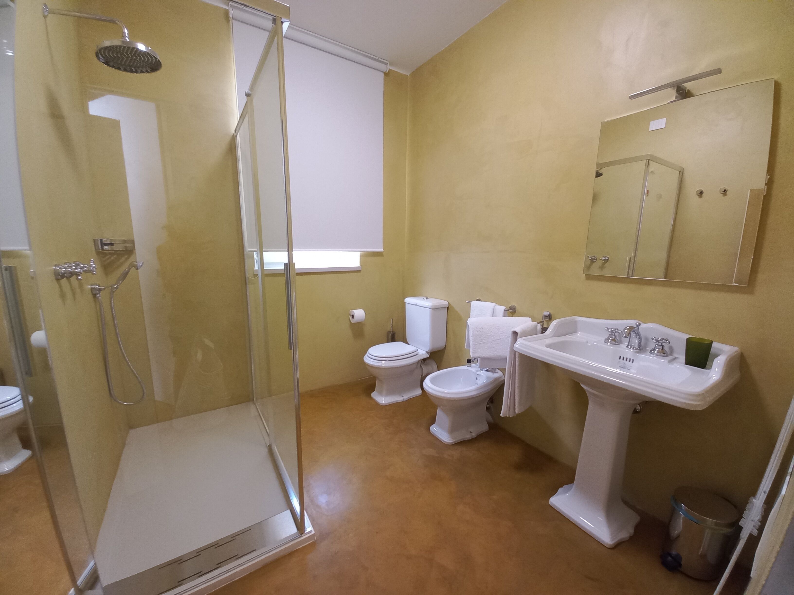 A bathroom with a glass-enclosed shower, a pedestal sink, two toilets, and a mirror above the sink.