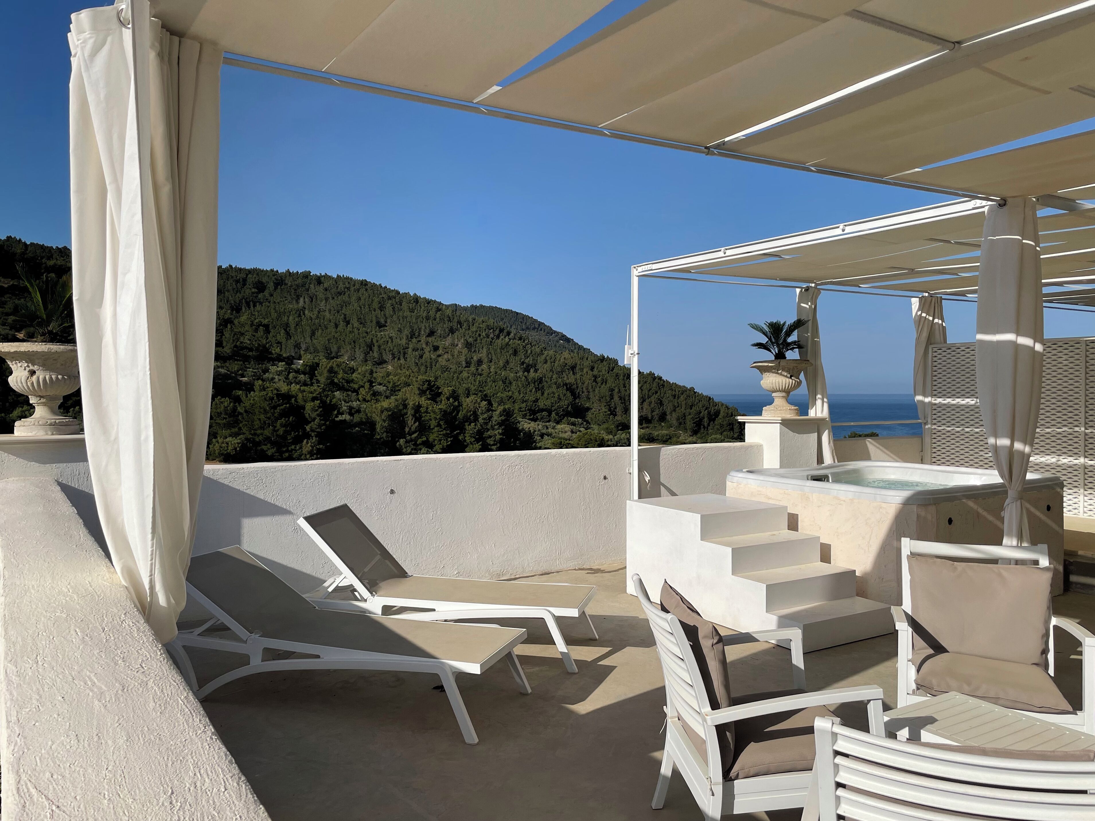 Una terraza con sillones blancos y un jacuzzi, con vistas a una ladera boscosa y al mar.