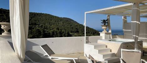 Una terraza con sillones blancos y un jacuzzi, con vistas a una ladera boscosa y al mar.