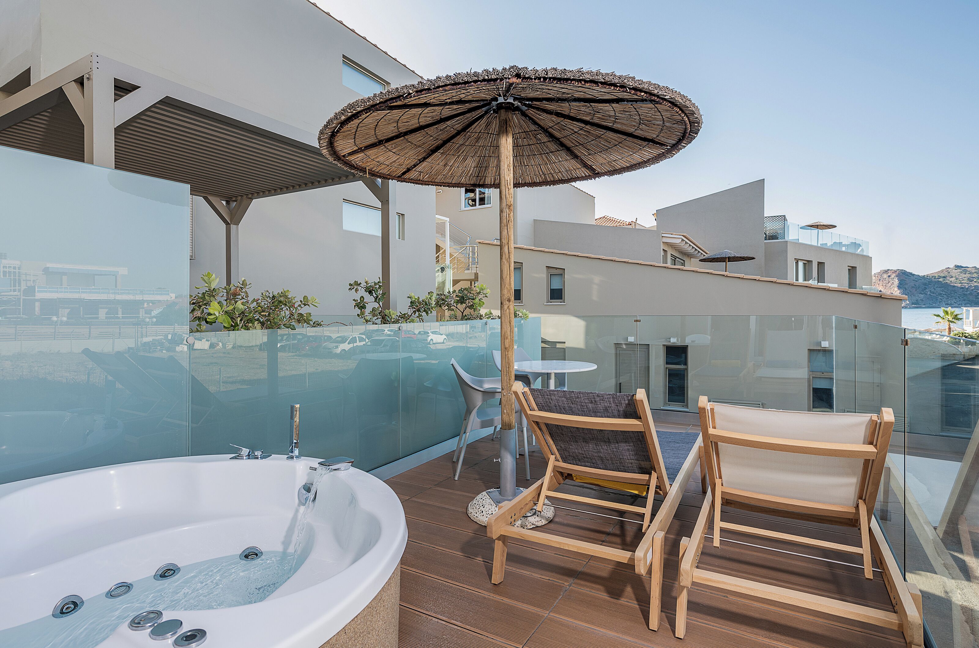 A modern outdoor balcony with a hot tub, wooden chairs, and a glass balustrade overlooking a scenic view.