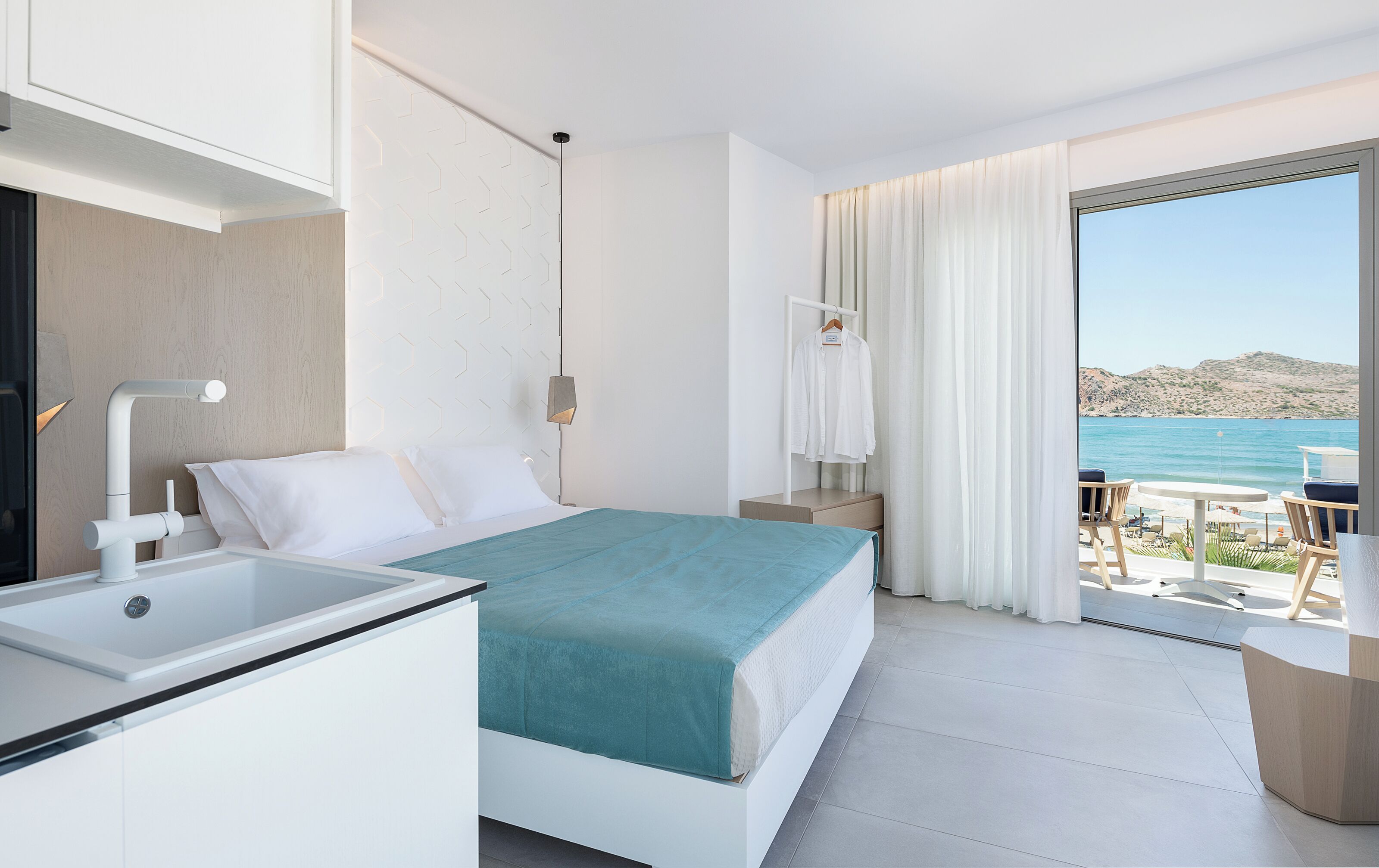 A modern hotel room with a bed, a desk, a view of the sea, and a balcony with outdoor seating.