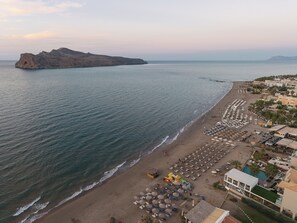 A beach with a row of sunbeds and umbrellas, a pool area, and a rocky outcrop in the distance. - Vergina Beach Resort (Chania)