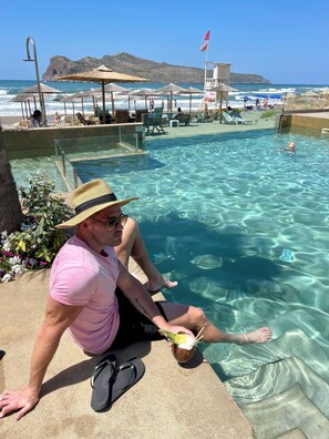 Een man zit aan een zwembad met helder water, met in de verte een strand met parasols en ligbedden.
