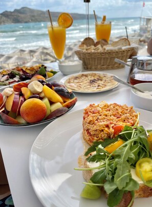 A table with breakfast items, including fruit, eggs, and bread, set on a beach with a view of the ocean. - Vergina Beach Resort (Chania)