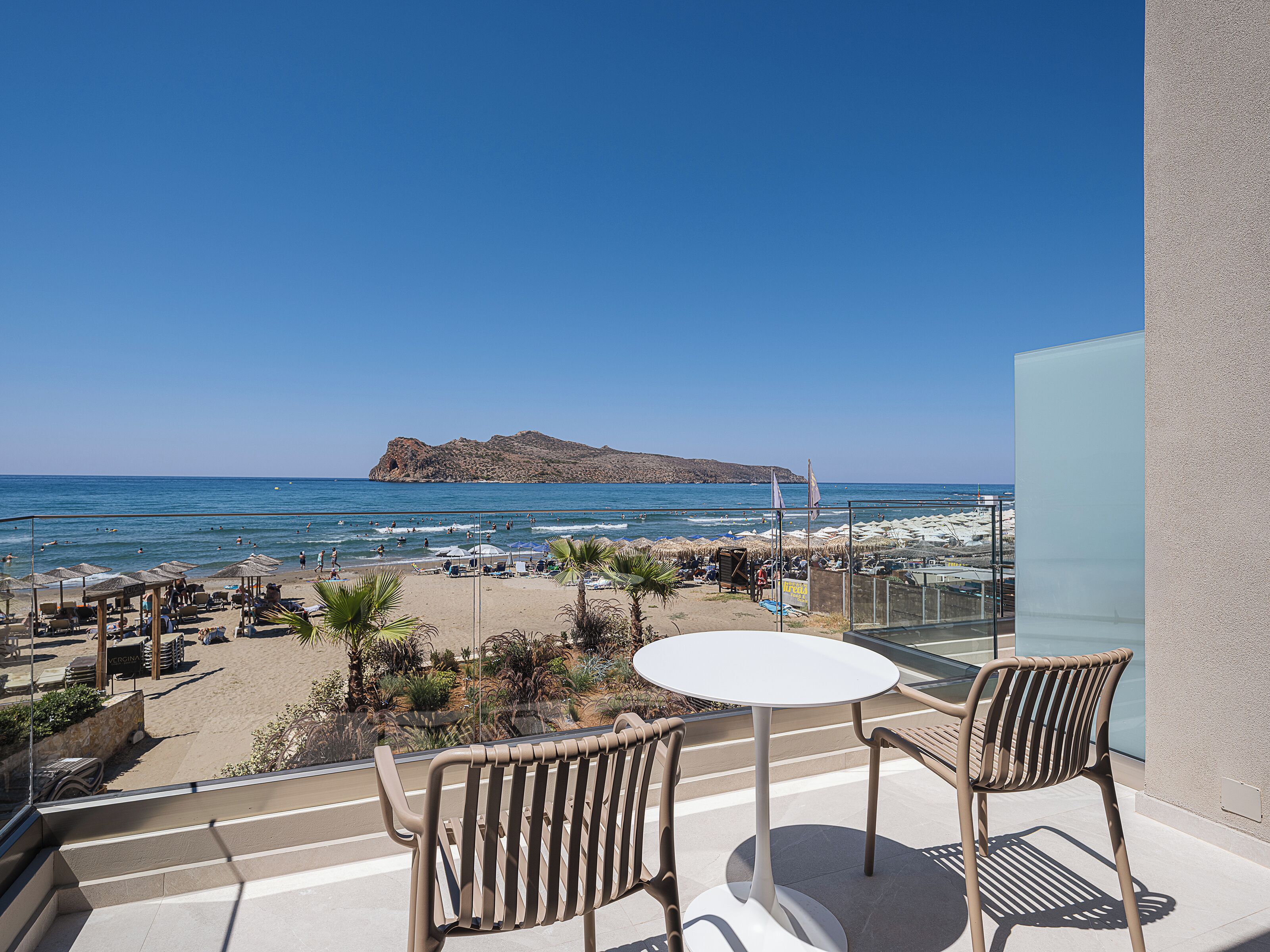 A balcony with a table and chairs overlooking a beach with umbrellas and lounge chairs.