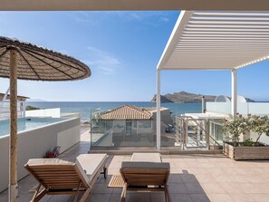 Un balcon avec vue sur la mer, des fauteuils de jardin en bois, une balustrade en verre et une pergola.