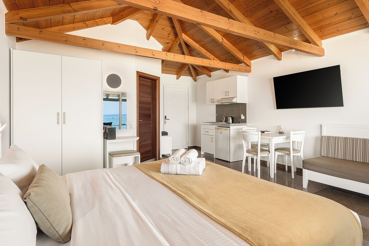 A bedroom with a large bed, a desk, a chair, a sofa, a TV, and a view of the sea.