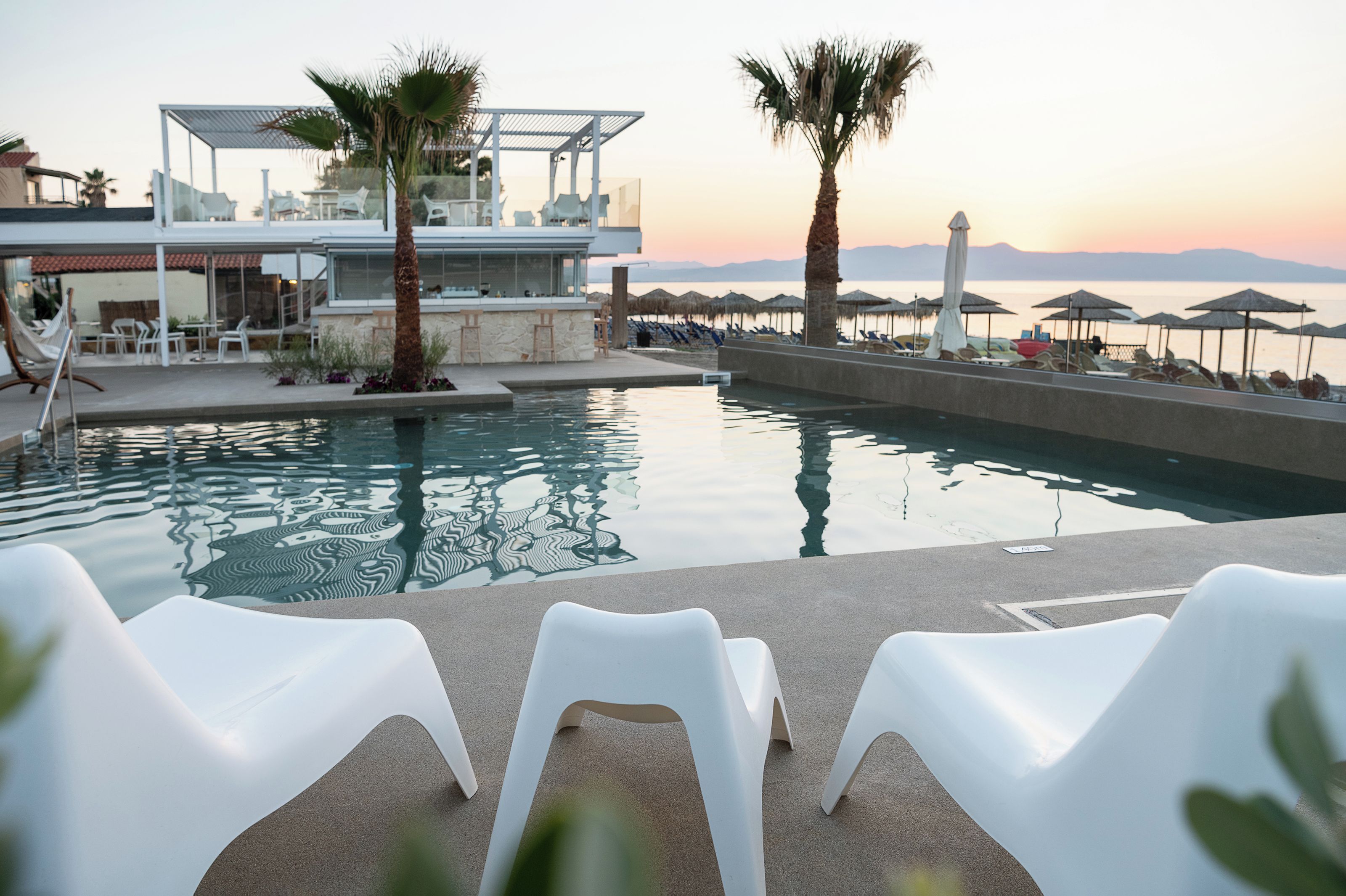 A pool area with lounge chairs, a bar, and a view of the sea at sunset.