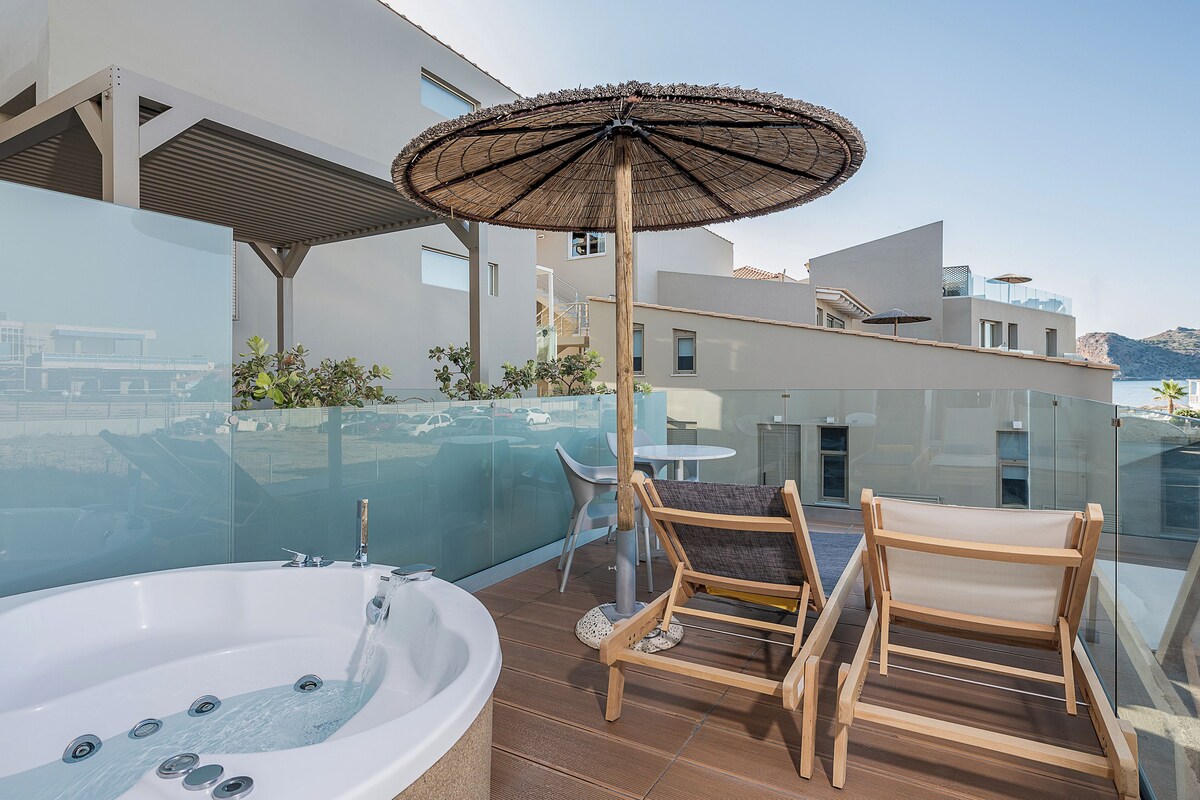 A modern outdoor balcony with a hot tub, wooden chairs, and a glass balustrade overlooking a scenic view.