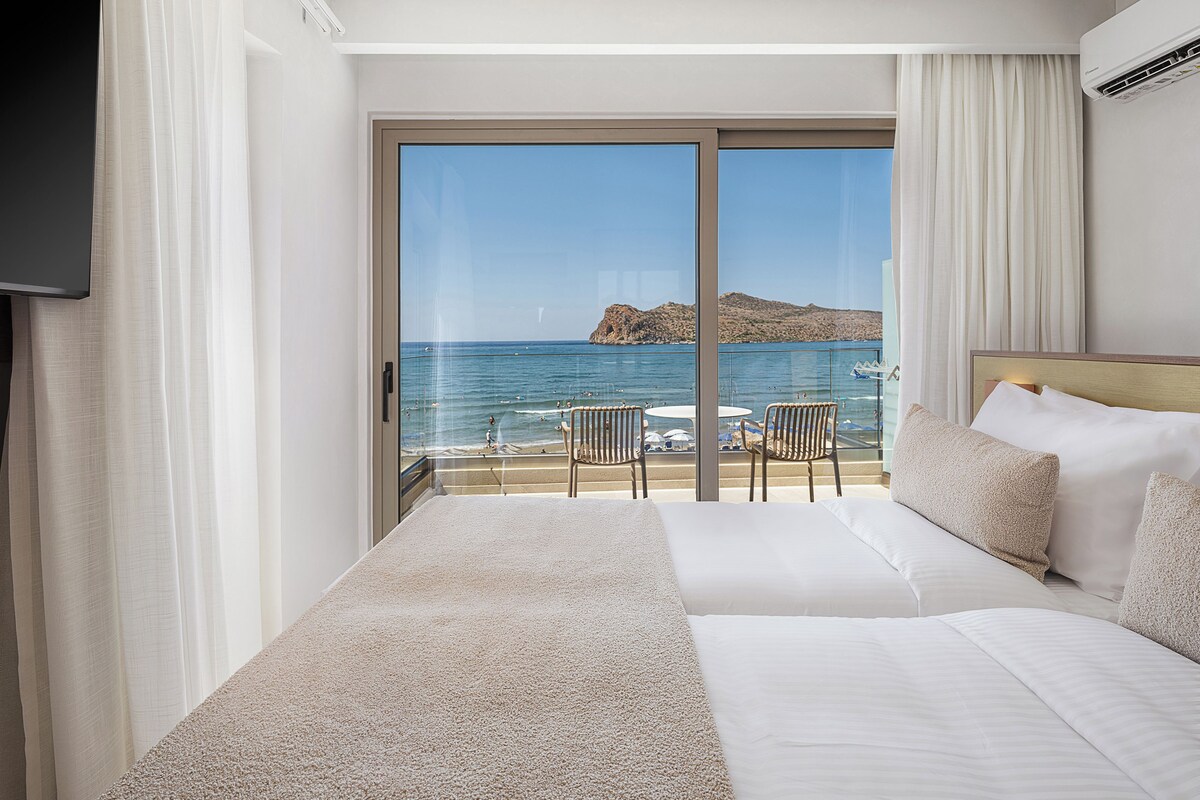A hotel room with a large bed, a TV, a balcony with a view of the sea, and a table with chairs.