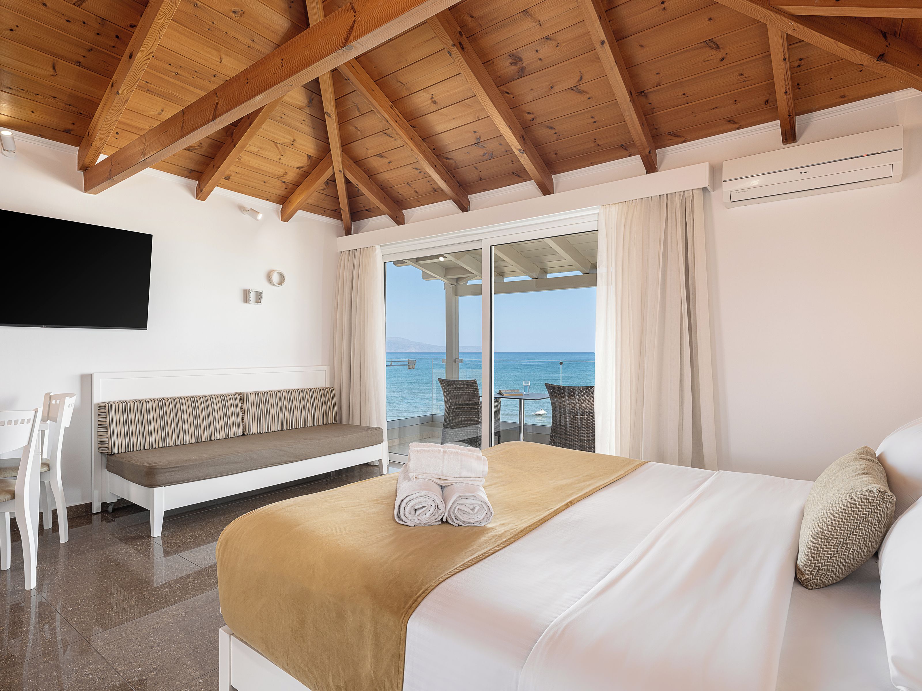 Habitación de hotel con techo de madera, cama con colcha color beige, sofá blanco, televisor montado en la pared y vista al mar a través de una puerta de cristal.
