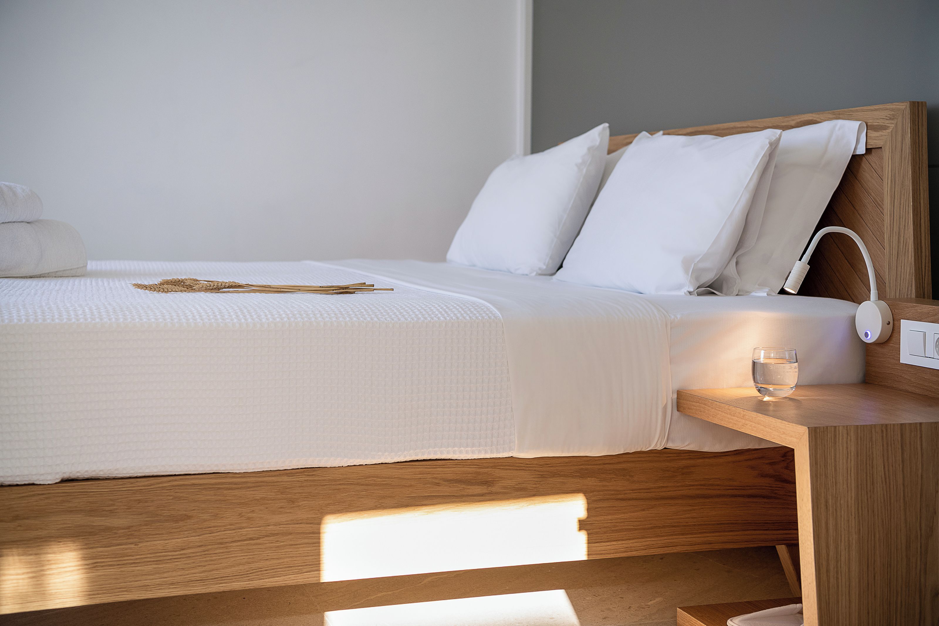 Un lit bien fait, avec du linge de lit blanc, une table de chevet sur laquelle se trouve un verre d’eau et une tête de lit en bois.