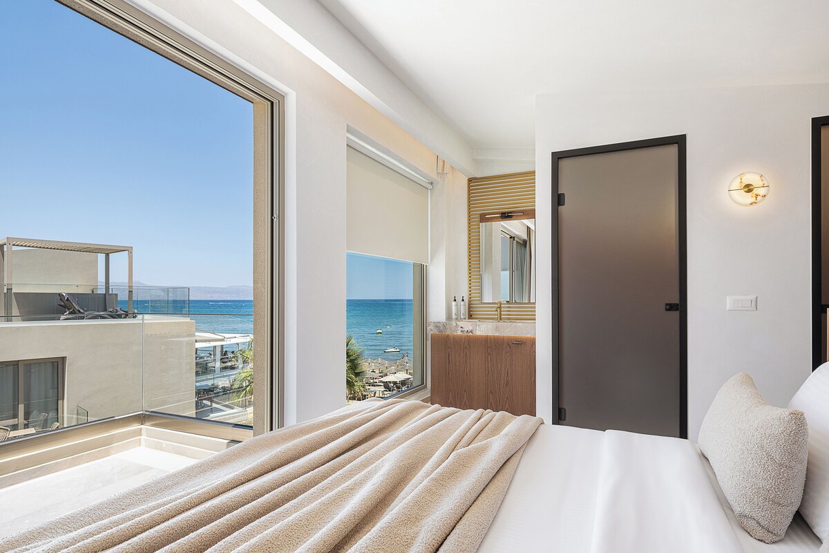 A bedroom with a large window, a bed, a bathroom with a mirror, and a view of the sea.